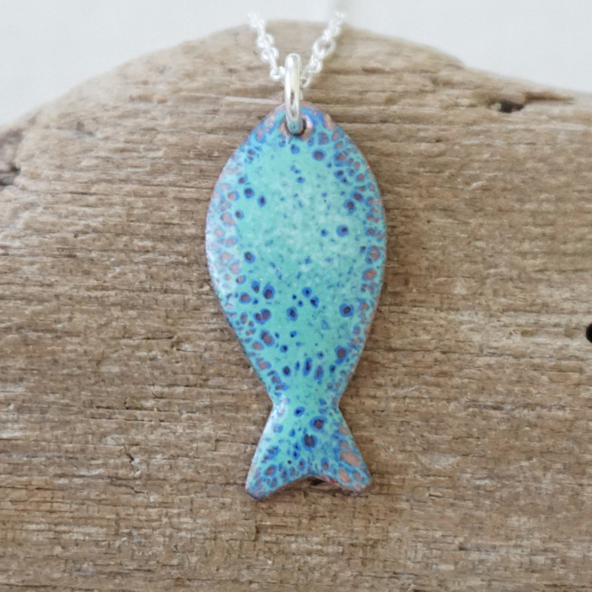 green and blue enamel fish shaped pendant against a wooden background