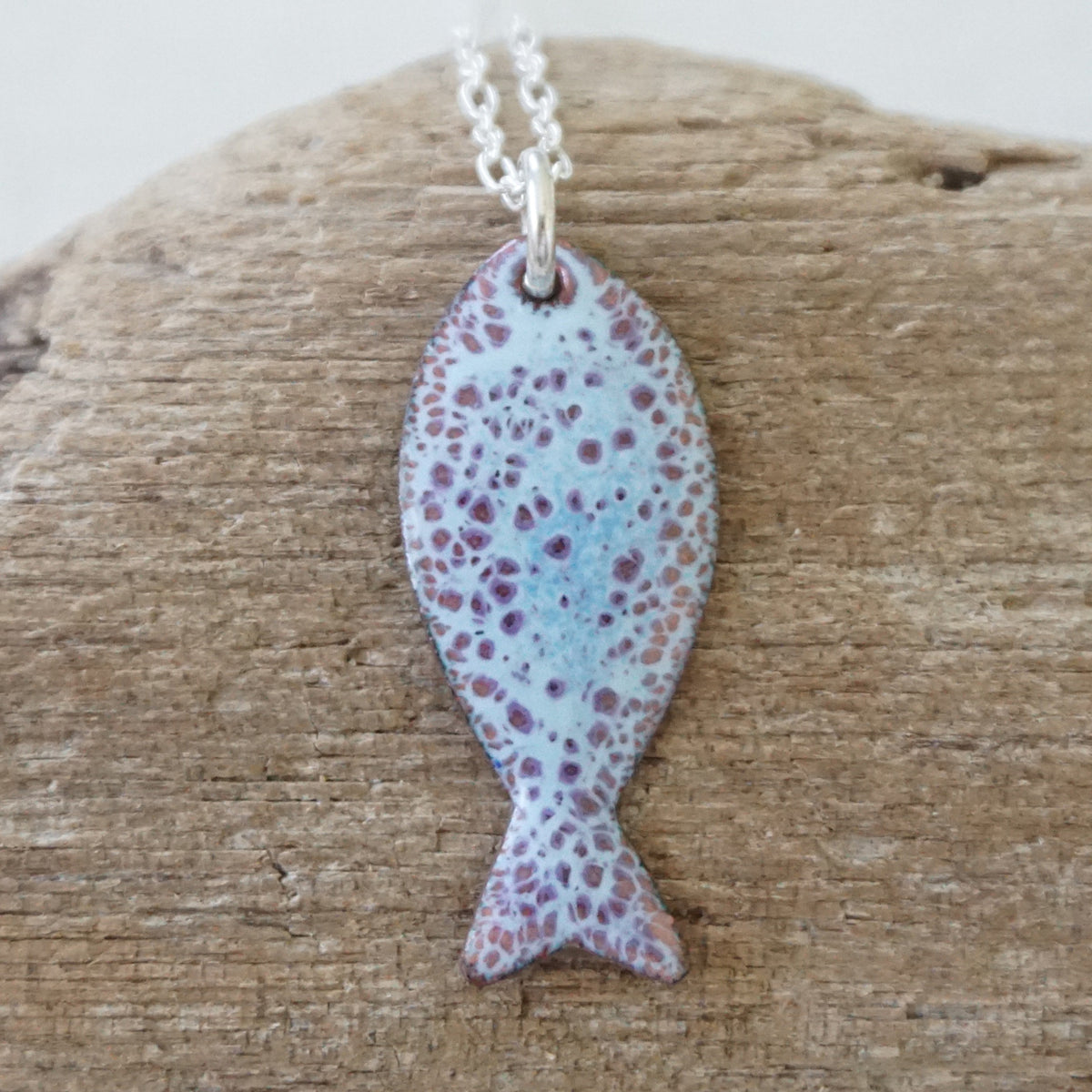 light blue and purple enamel fish shaped pendant against a wooden background