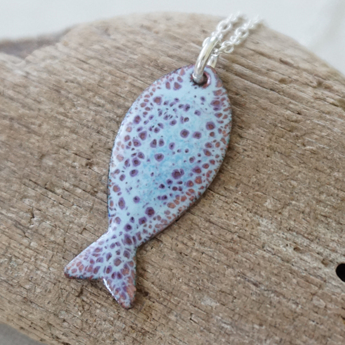 light blue and purple enamel fish shaped pendant against a wooden background