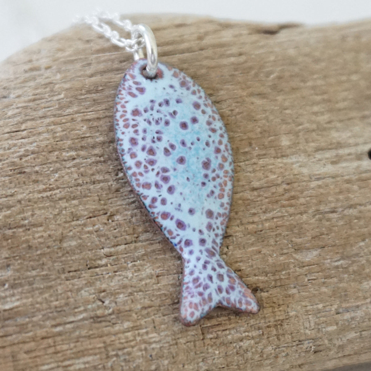 light blue and purple enamel fish shaped pendant against a wooden background