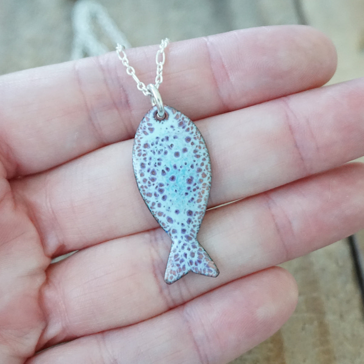 hand holding a light blue and purple enamel fish shaped pendant against a wooden background