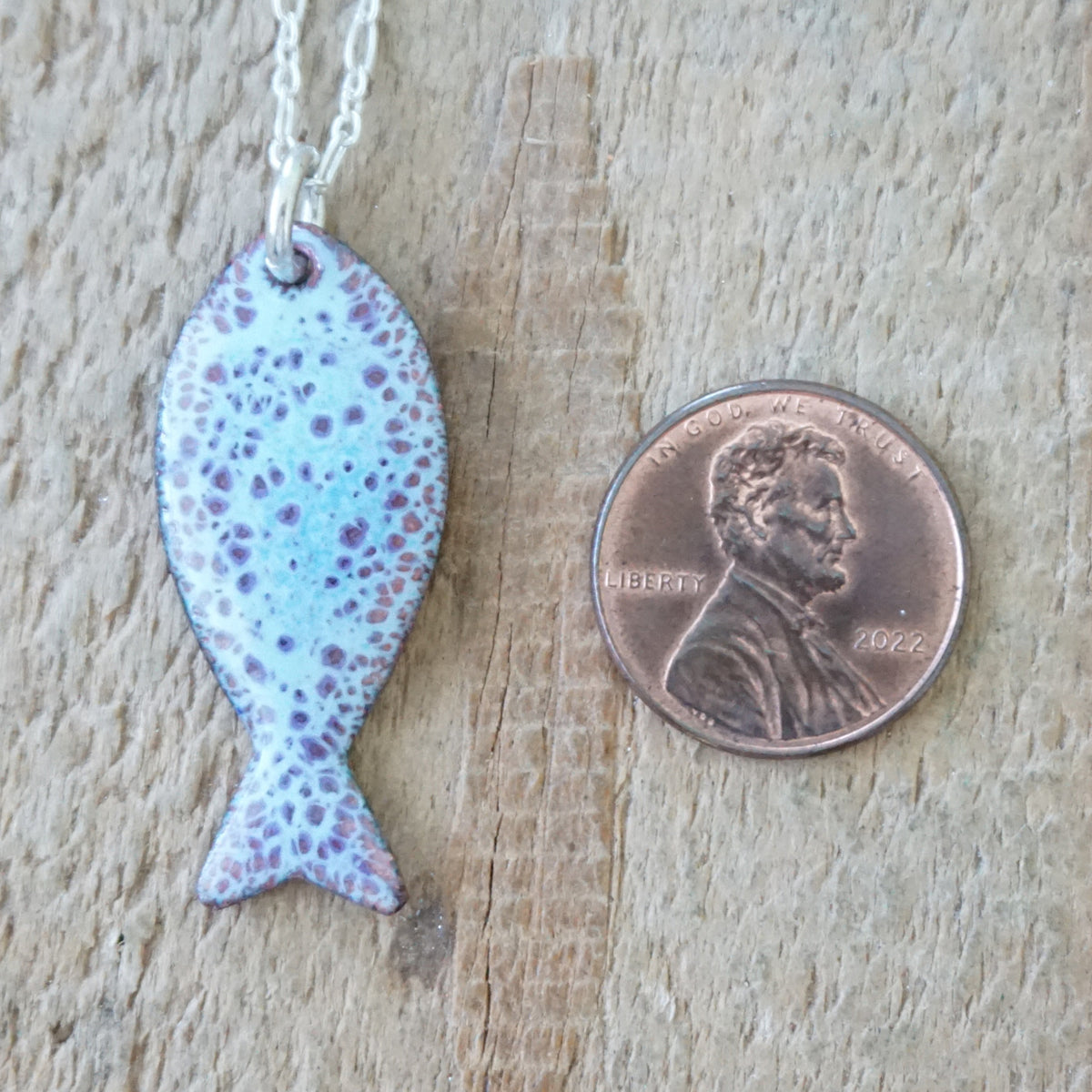 light blue and purple enamel fish shaped pendant, next to a penny for scale, against a wooden background