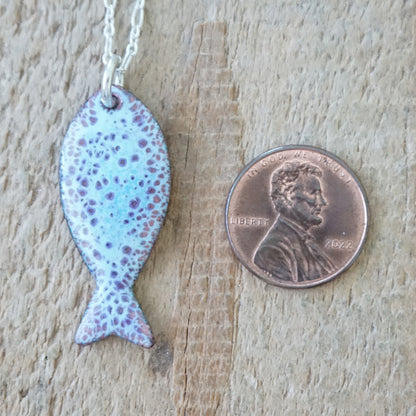 light blue and purple enamel fish shaped pendant, next to a penny for scale, against a wooden background