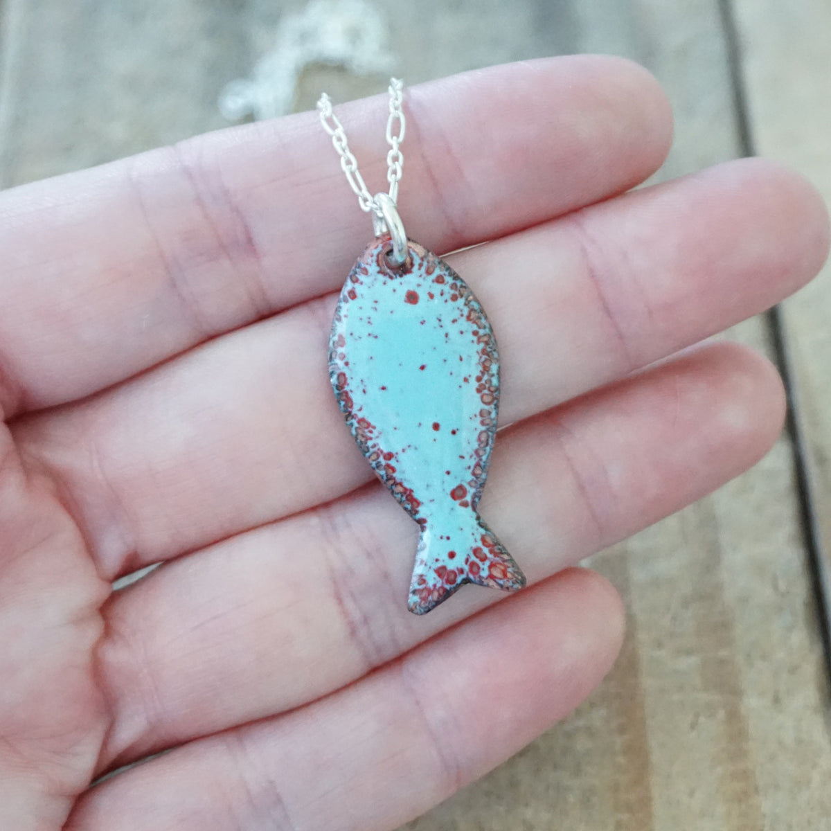 hand holding a light blue and red enamel fish shaped pendant against a wooden background