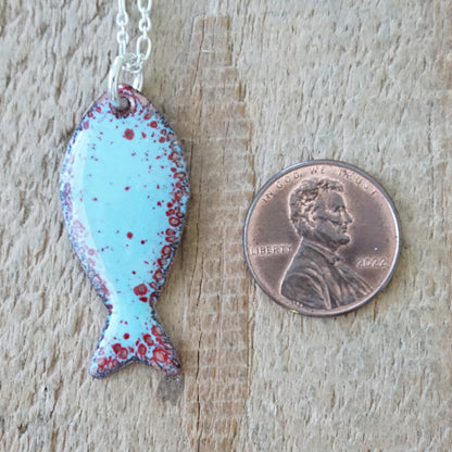 light blue and red enamel fish shaped pendant, next to a penny for scale, against a wooden background