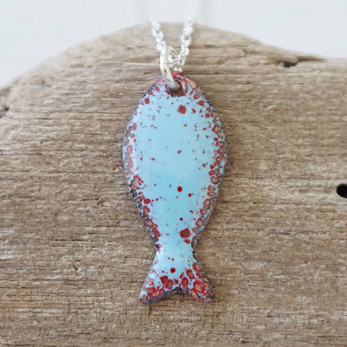 light blue and red enamel fish shaped pendant against a wooden background