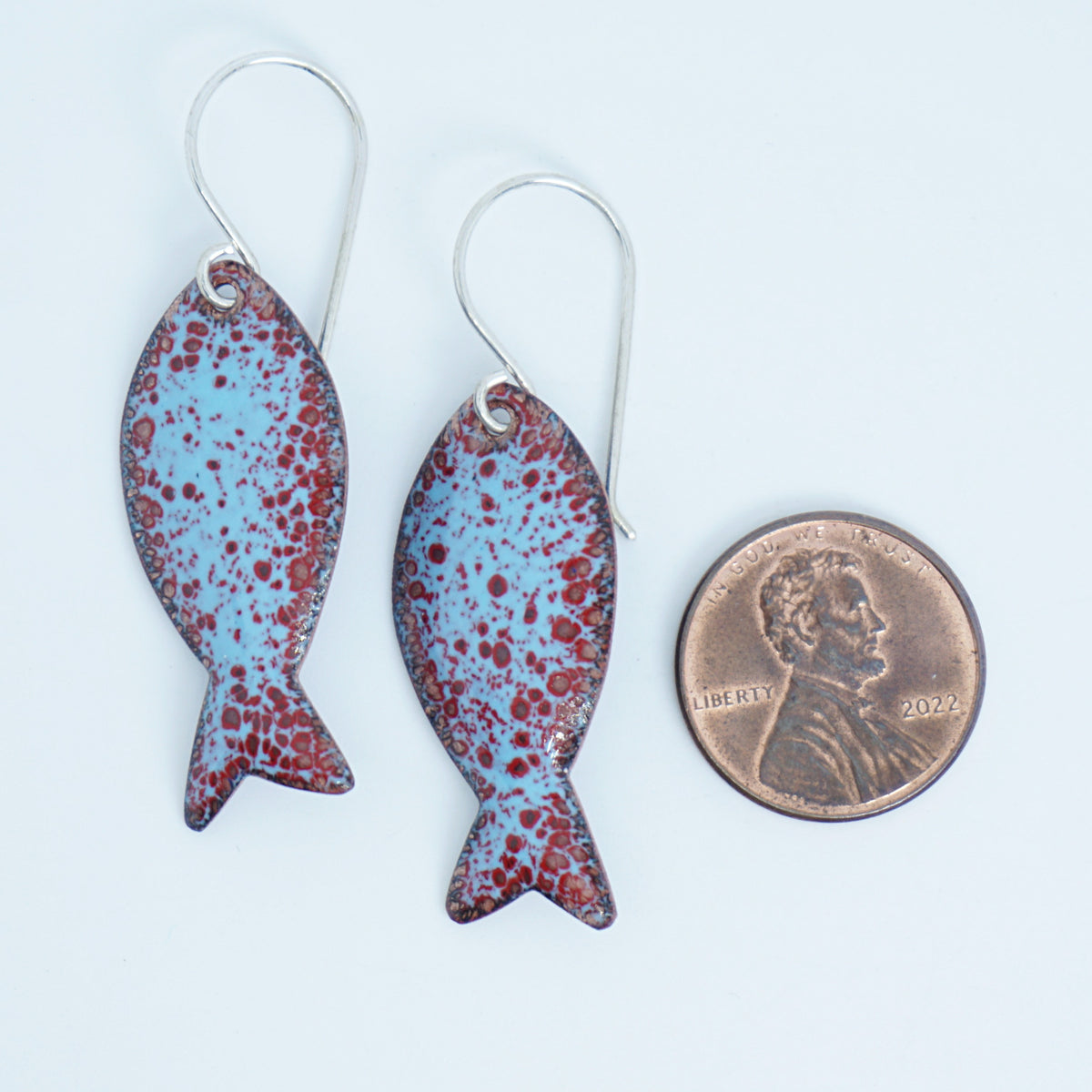 light blue and red enamel fish shaped earrings, next to a penny for scale, against a white background