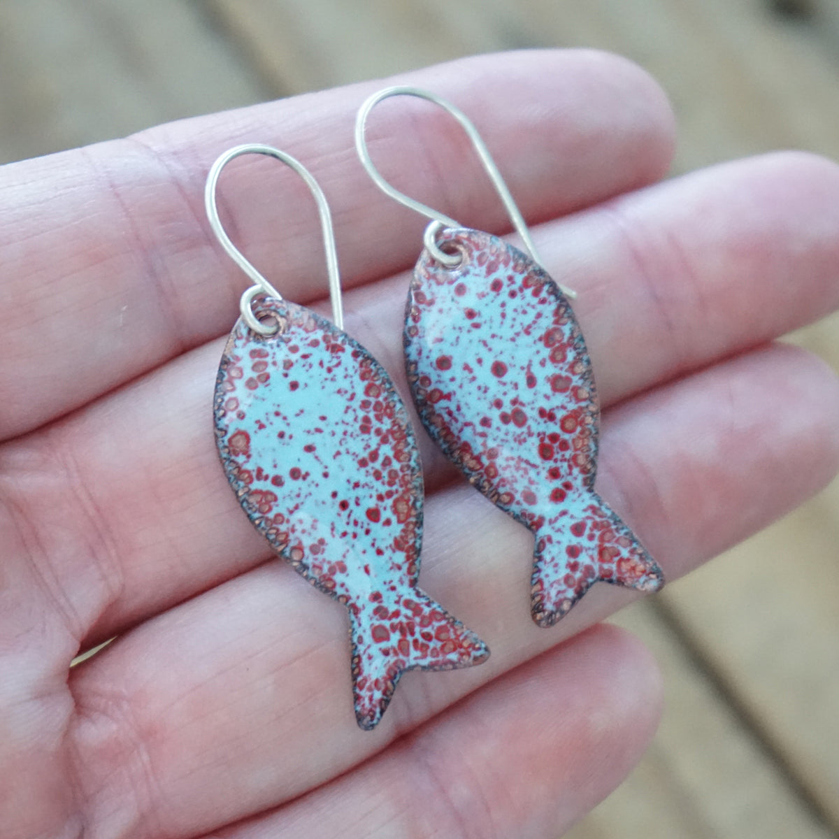 hand holding a pair of light blue and red enamel fish shaped earrings, against a wooden background