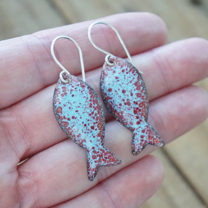 hand holding a pair of light blue and red enamel fish shaped earrings, against a wooden background