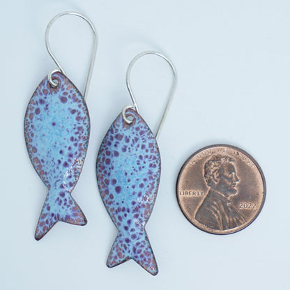 light blue and purple enamel fish shaped earrings, next to a penny for scale, against a white background