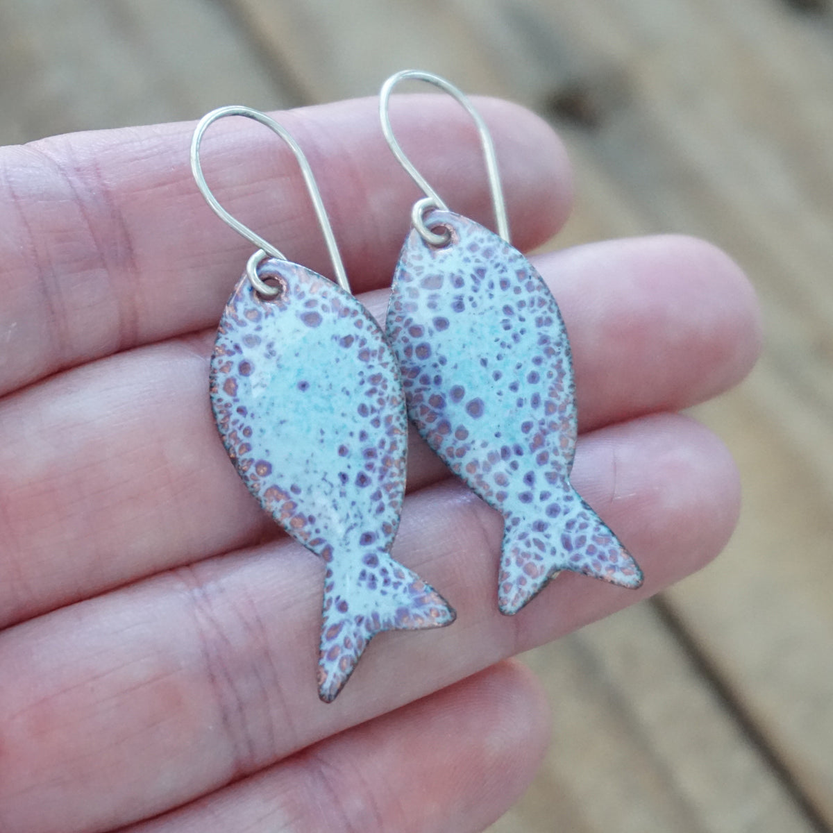 hand holding a light blue and purple enamel fish shaped earrings, against a wooden background