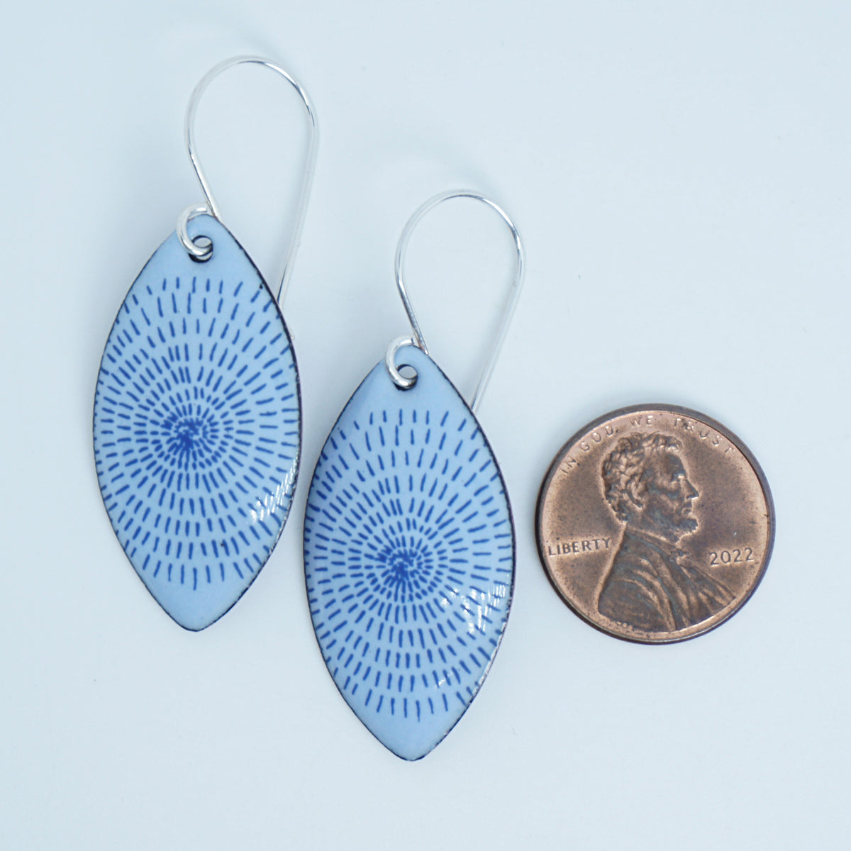 a pair of light purple enamel marquis shaped earrings with blue accents, next to a penny for scale, against a white background