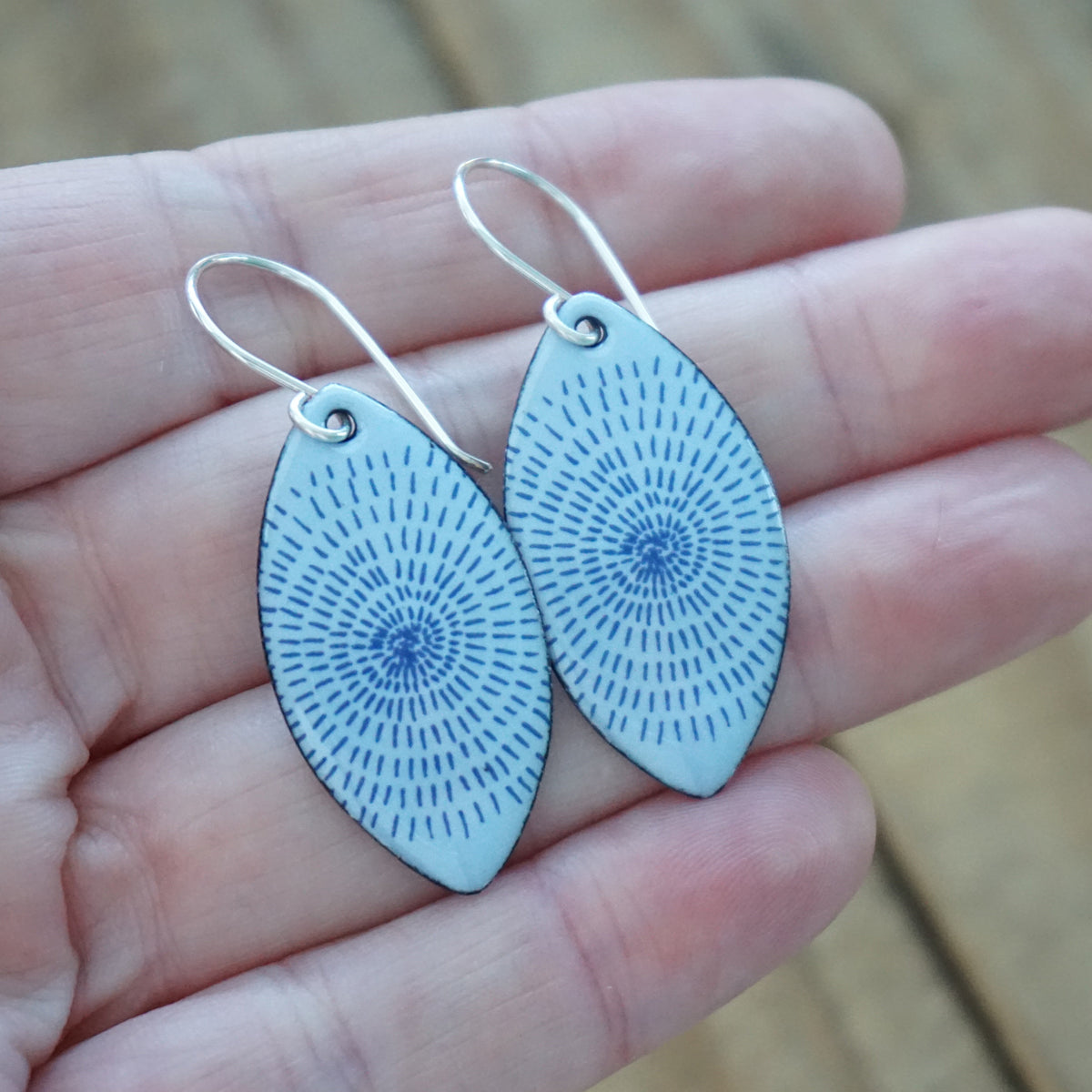 hand holding a pair of light purple enamel marquis shaped earrings with blue accents, against a wooden background