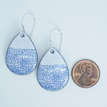 a pair of white enamel teardrop shaped earrings with blue accents, next to a penny for scale, against a white background