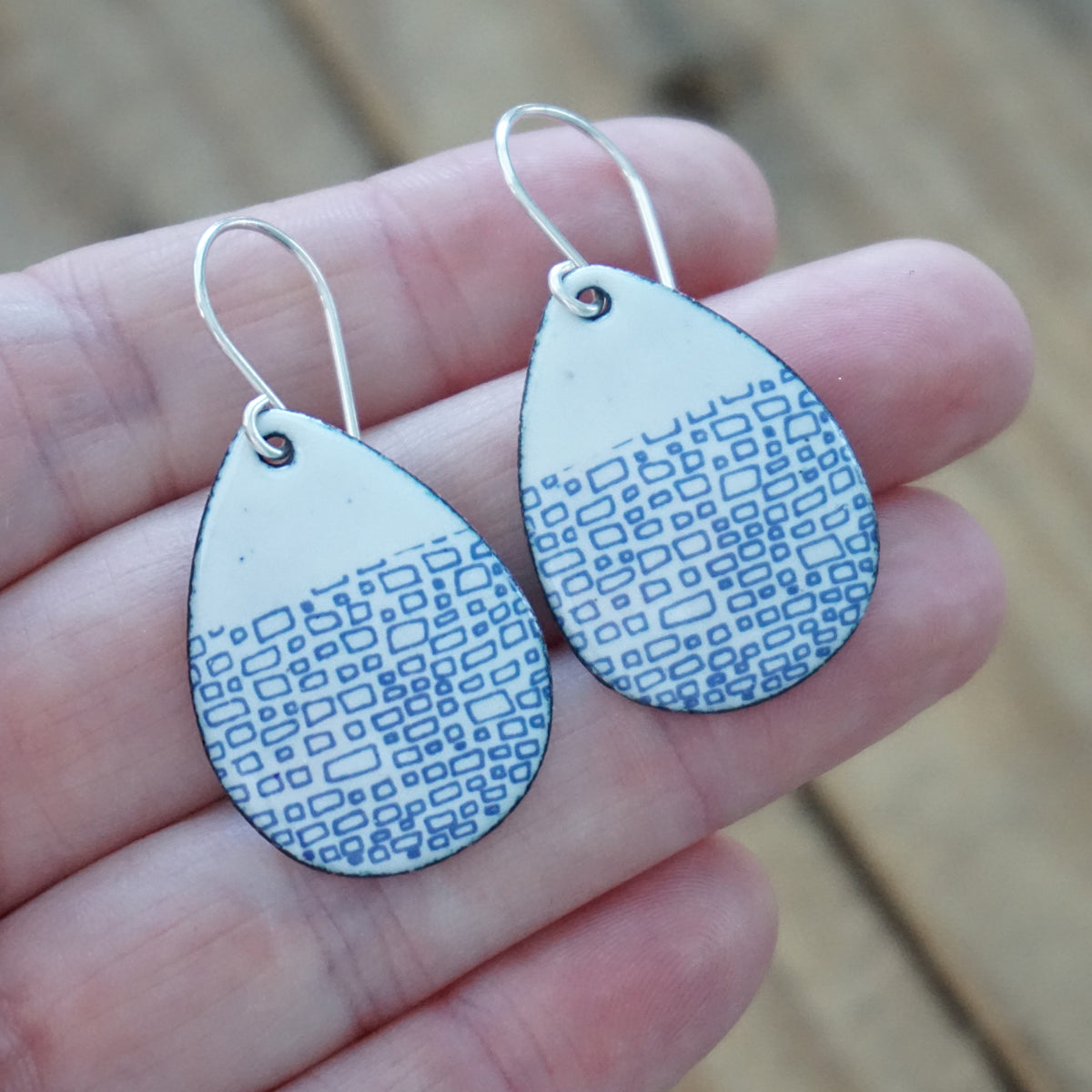 hand holding a pair of white enamel teardrop shaped earrings with blue accents, against a wooden background