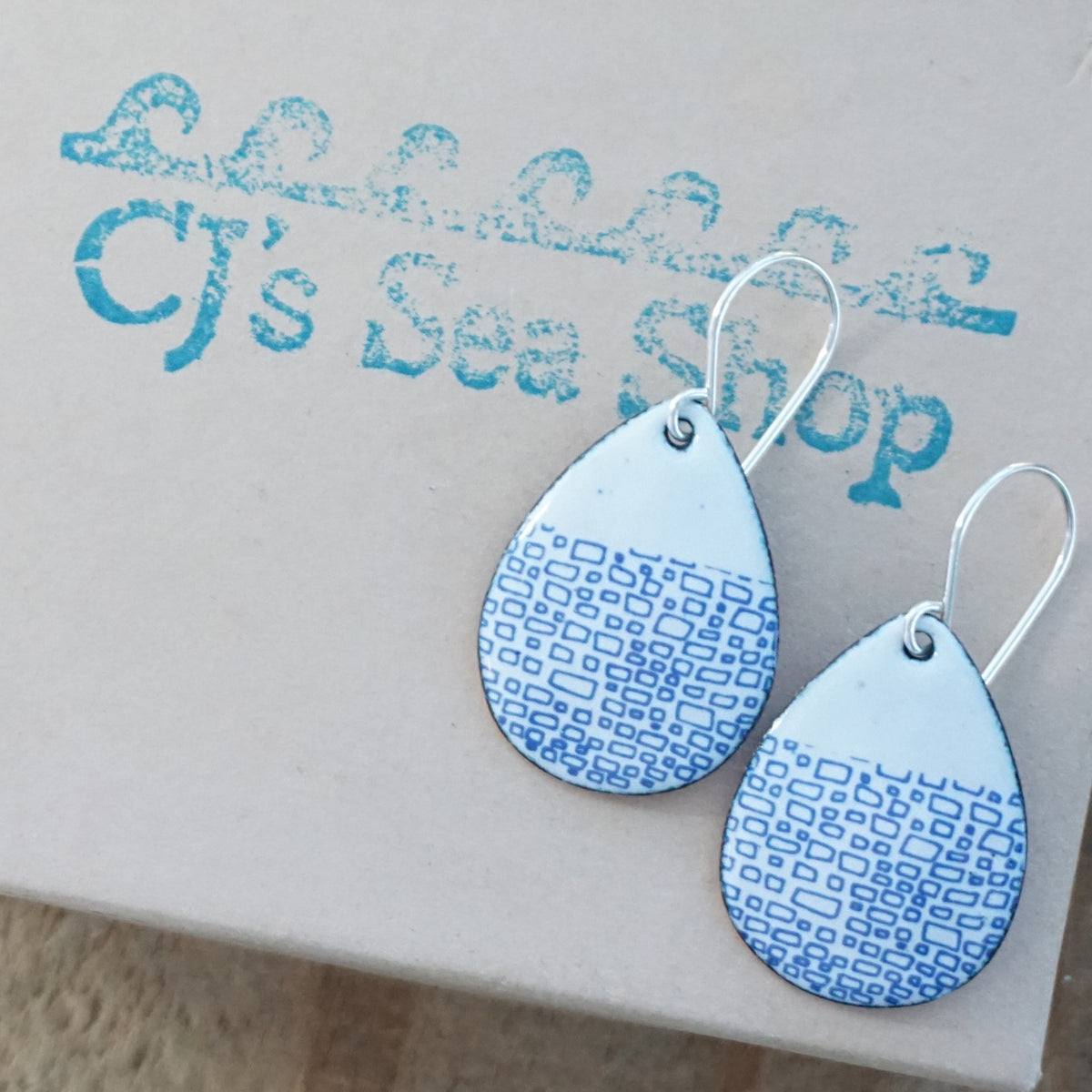 a pair of white enamel teardrop shaped earrings with blue accents, displayed on a gift box