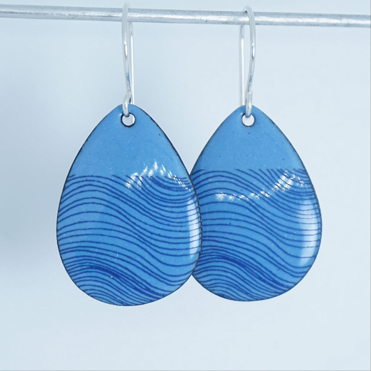 a pair of blue enamel teardrop shaped earrings with blue wavy line accents, against a white background
