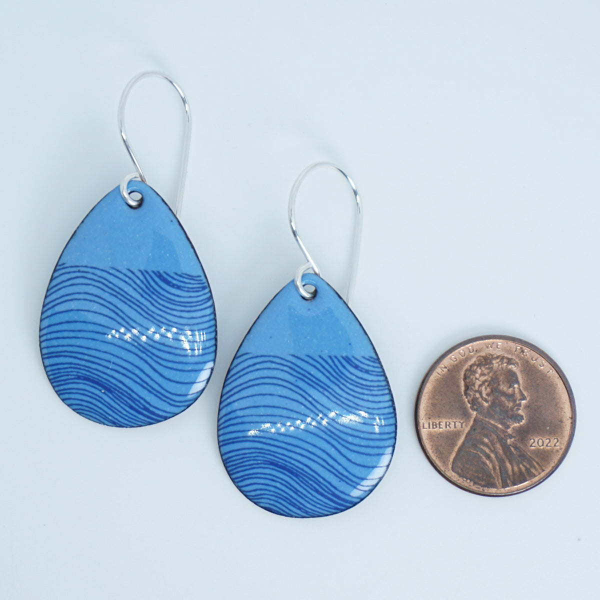 a pair of blue enamel teardrop shaped earrings with blue wavy line accents, next to a penny for scale, against a white background