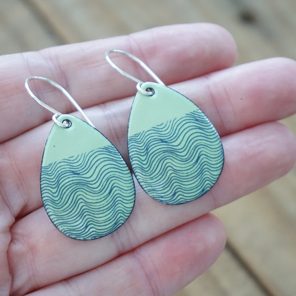 hand holding a pair of green enamel teardrop shaped earrings with blue wavy line accents, against a wooden background