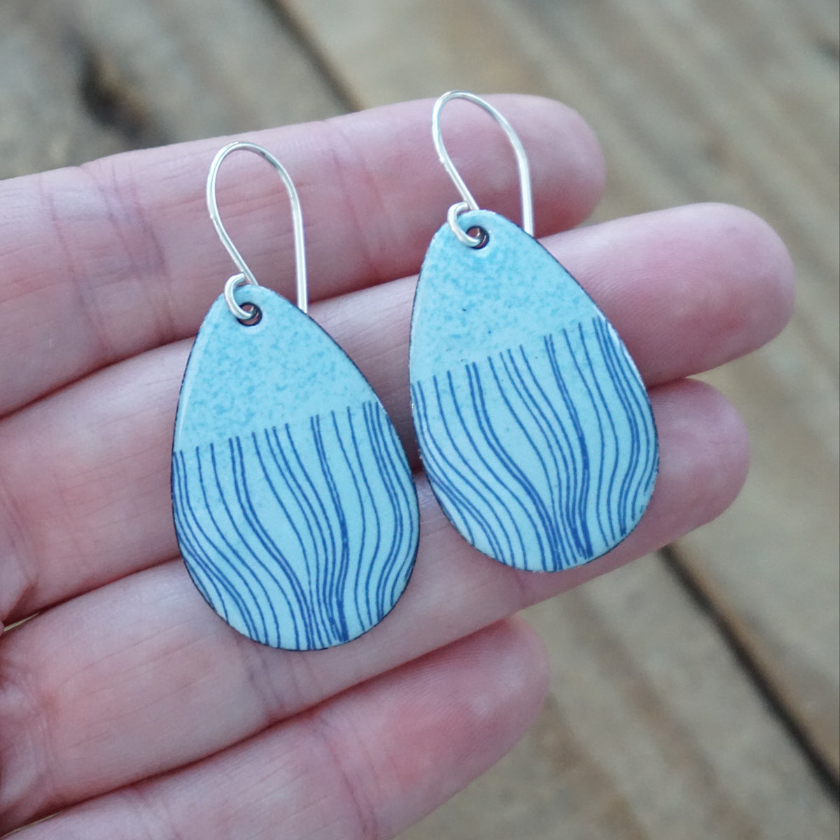 hand holding a pair of light blue enamel teardrop shaped earrings with blue wavy line accents, against a wooden background