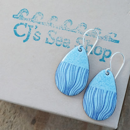 a pair of light blue enamel teardrop shaped earrings with blue wavy line accents, displayed on a gift box