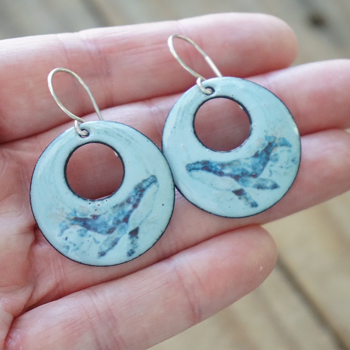 Round blue earrings with whale design held in a hand against a beige background