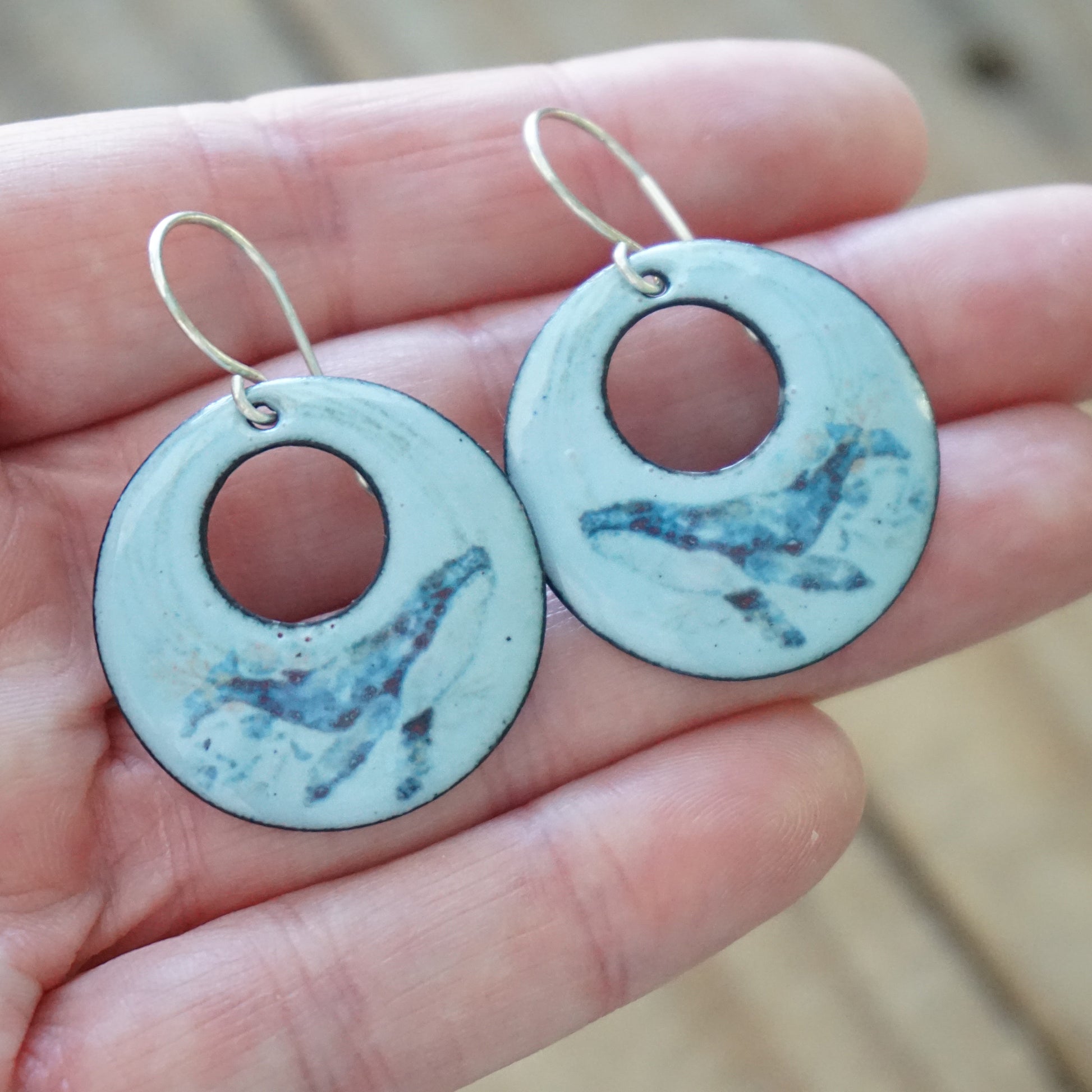 Round blue earrings with whale design held in a hand against a beige background
