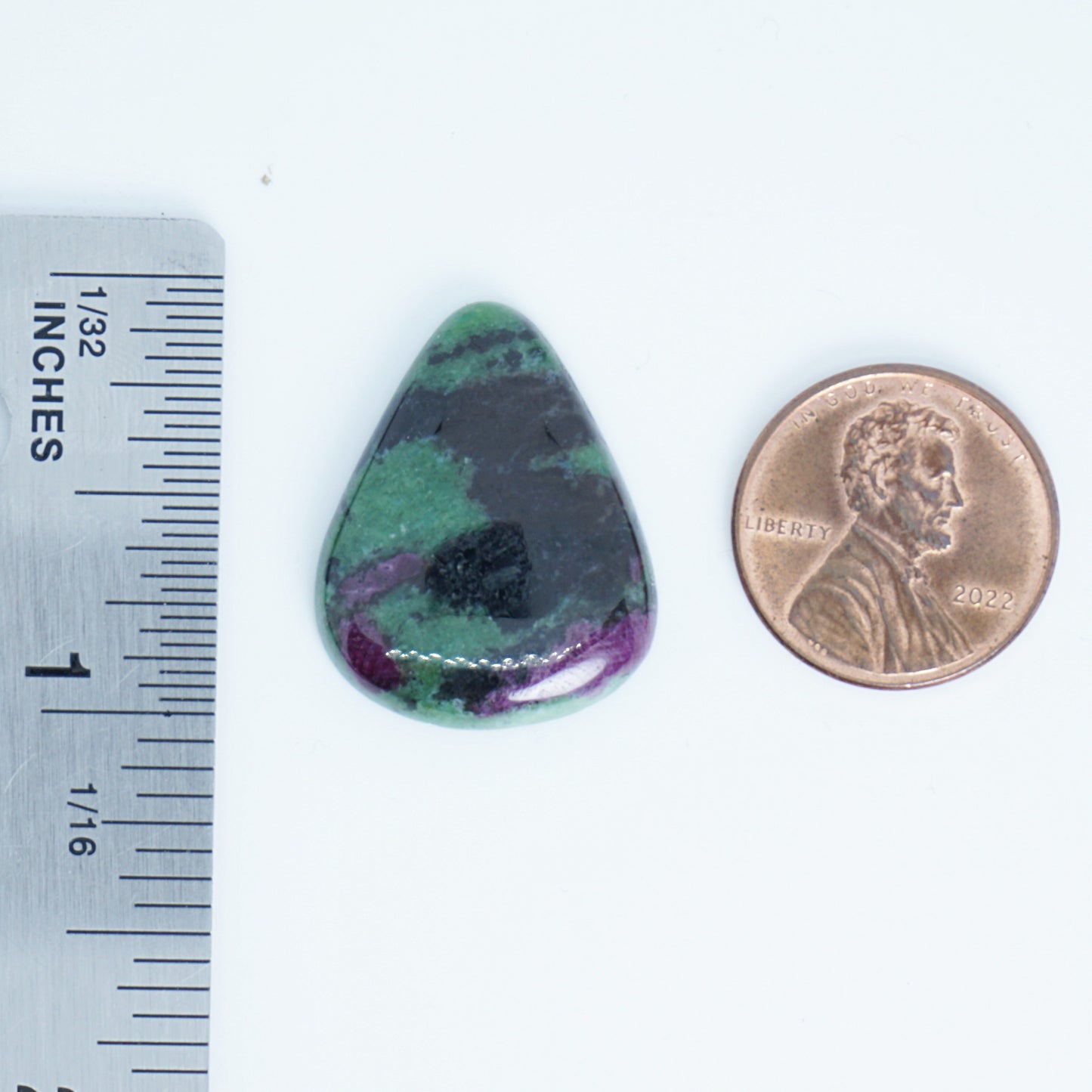 Teardrop-shaped green and purple stone next to a penny and ruler for scale on a white background