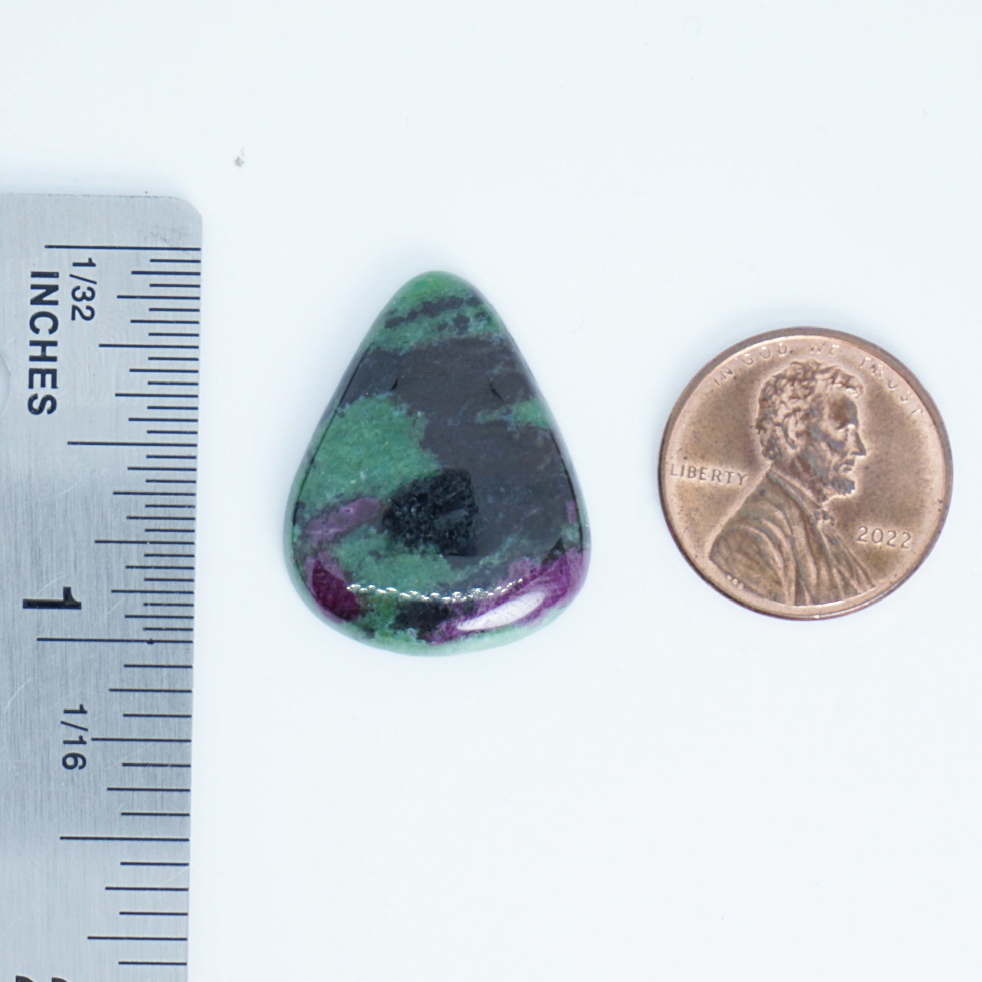 Teardrop-shaped green and purple stone next to a penny and ruler for scale on a white background