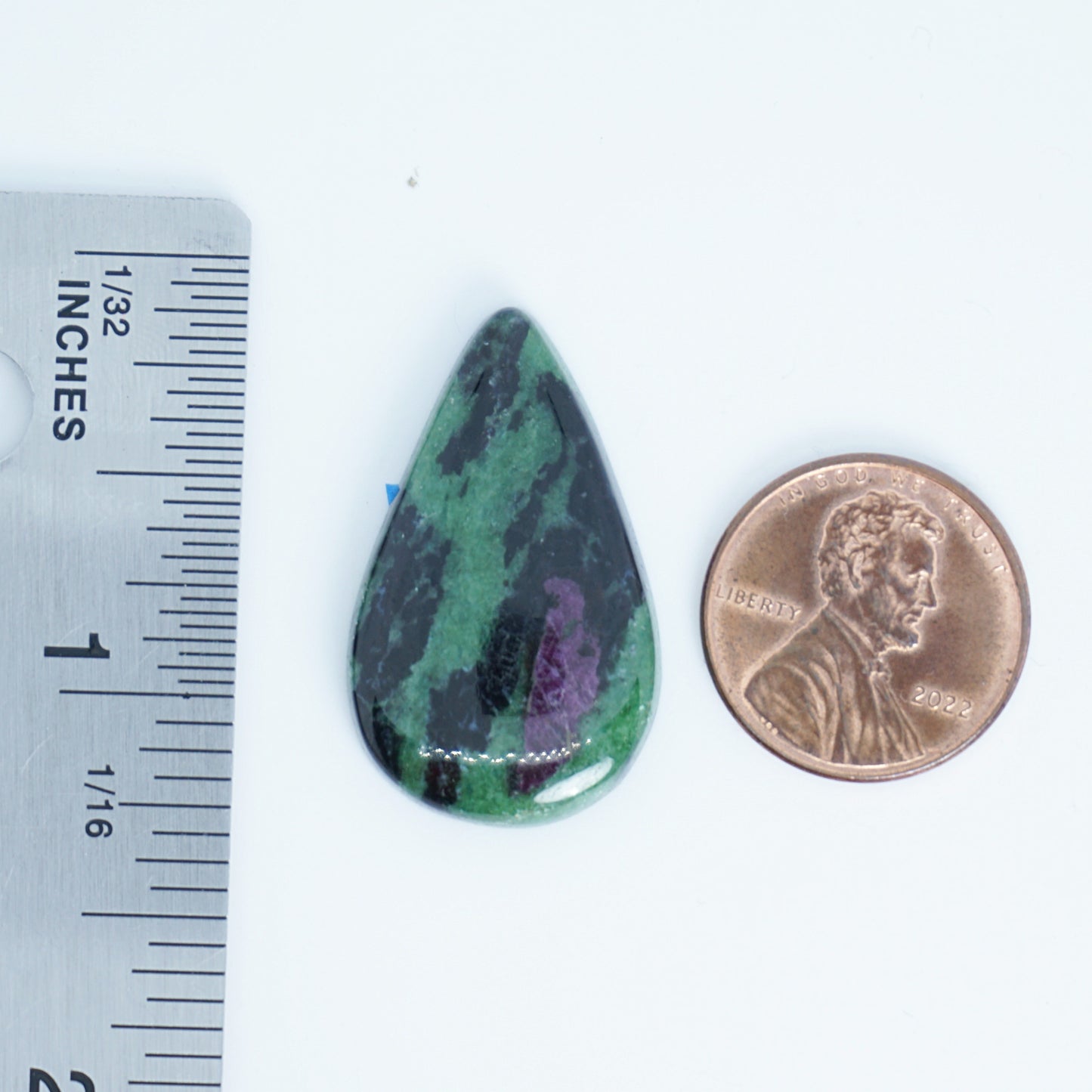 Teardrop-shaped green and black stone next to a penny and ruler for size comparison on a white background.
