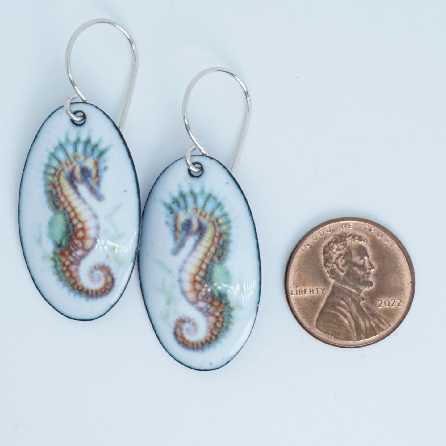 Oval earrings with seahorse design next to a penny for scale on a white background