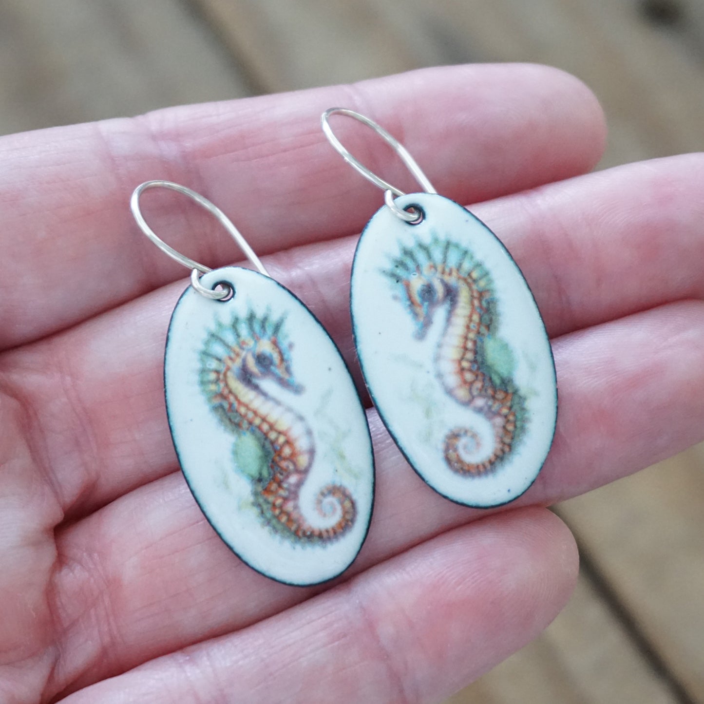Oval-shaped earrings with seahorse design held in a hand.