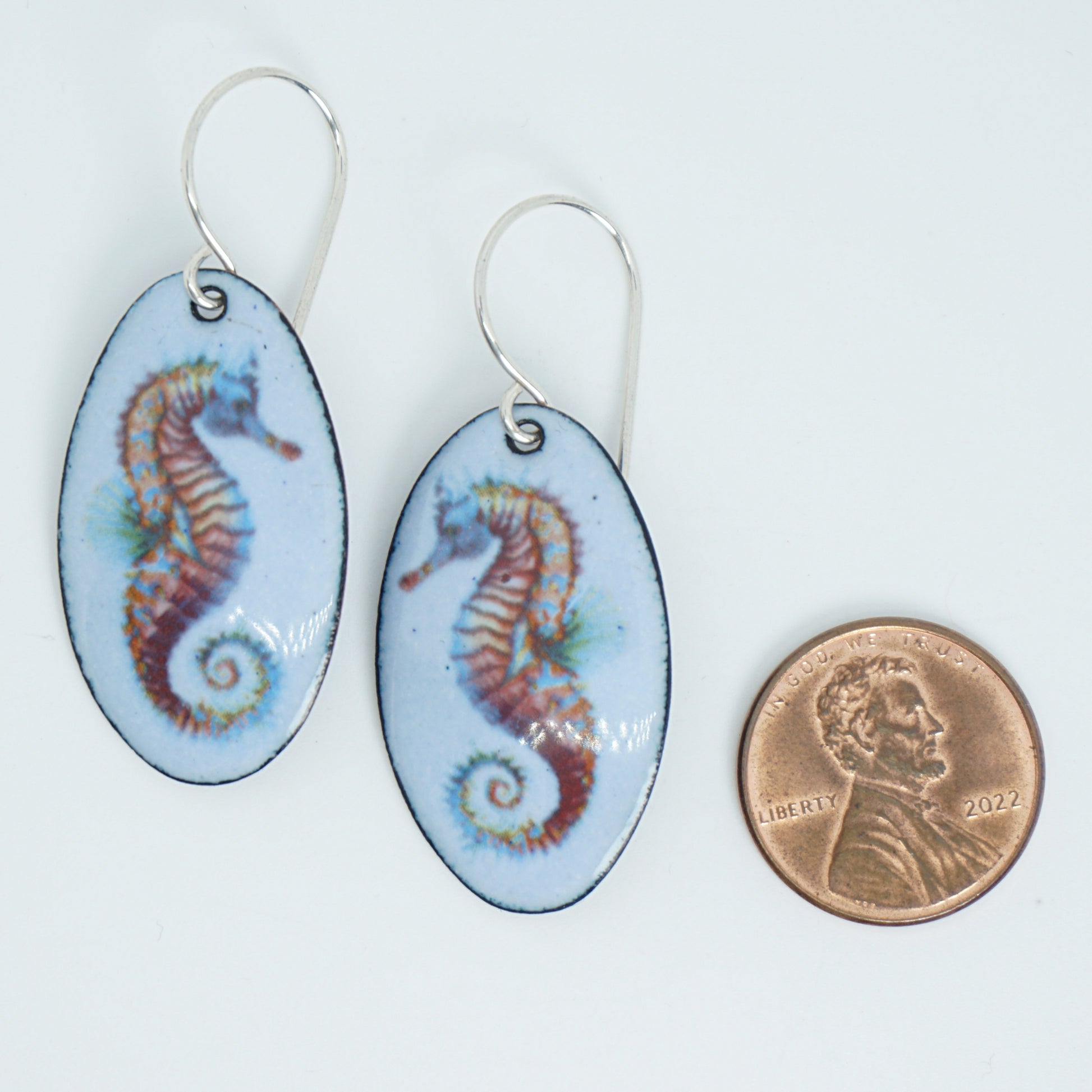 Oval-shaped earrings with seahorse design next to a penny for scale on a white background