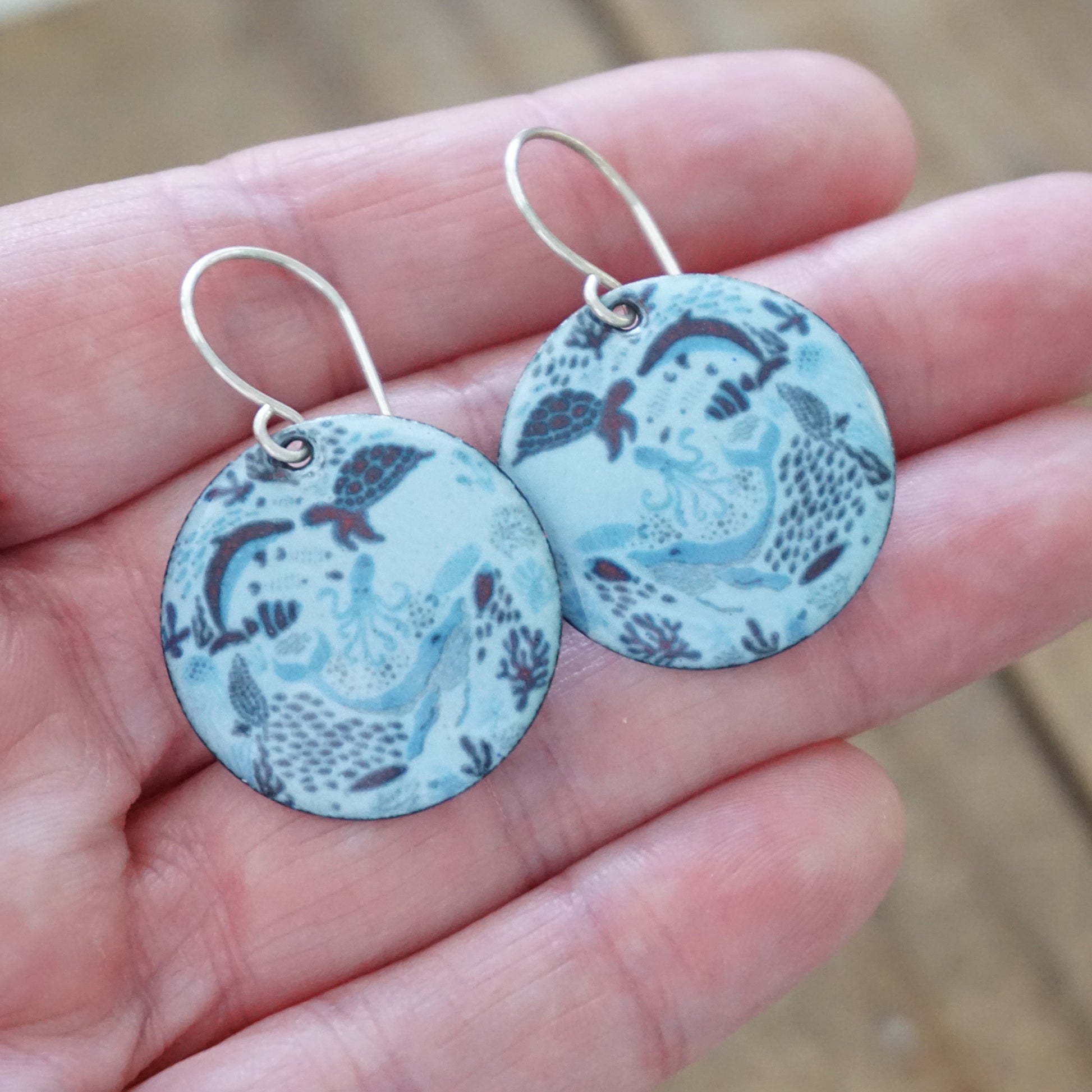Pair of blue sea life patterned earrings held in a hand with a blurred background