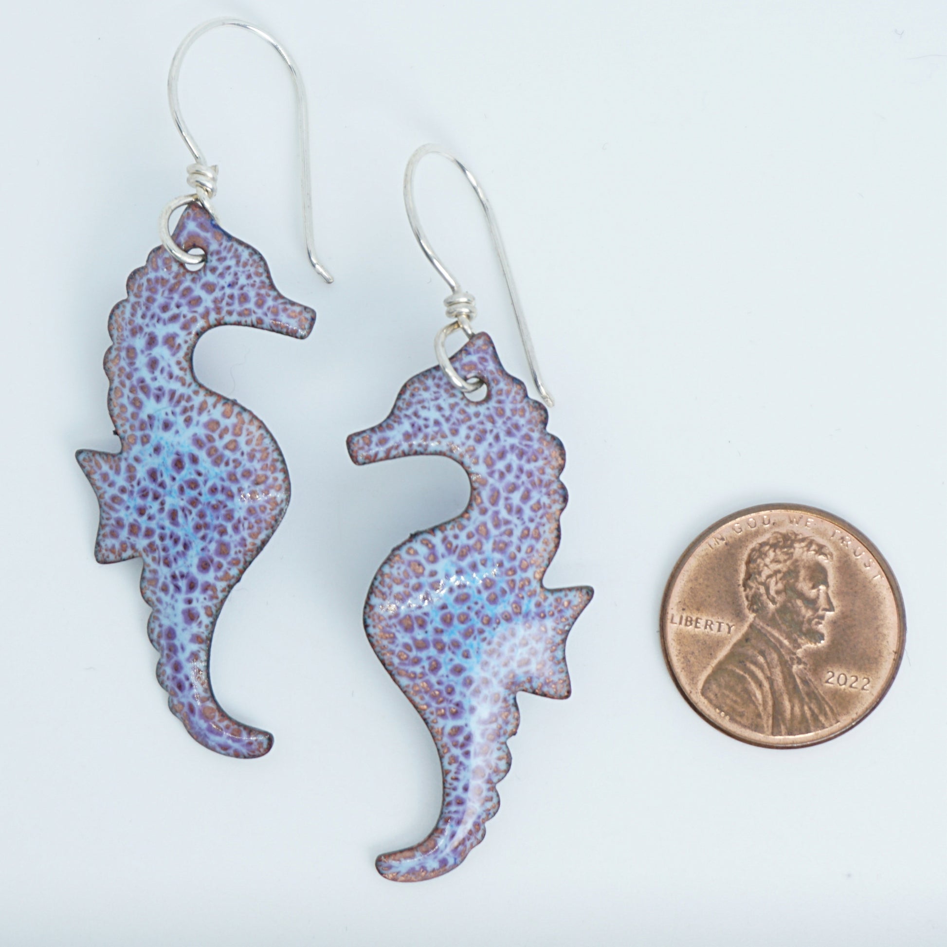Purple seahorse earrings next to a penny for scale on a white background