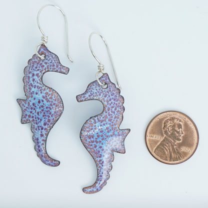 Purple seahorse earrings next to a penny for scale on a white background