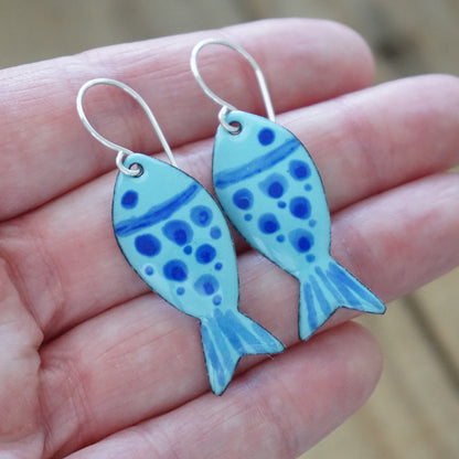 Blue fish-shaped earrings held in a hand with a blurred background