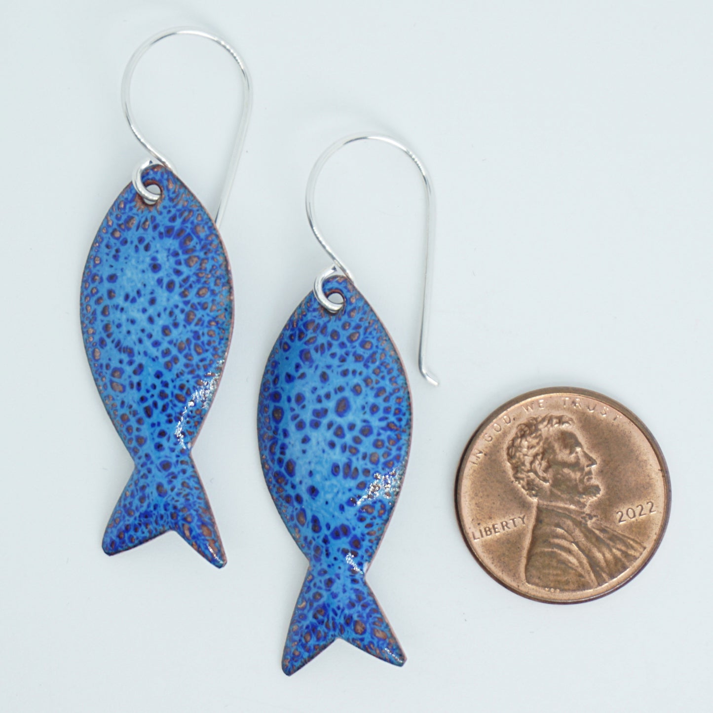 Blue speckled fish-shaped earrings next to a penny for scale on a white background