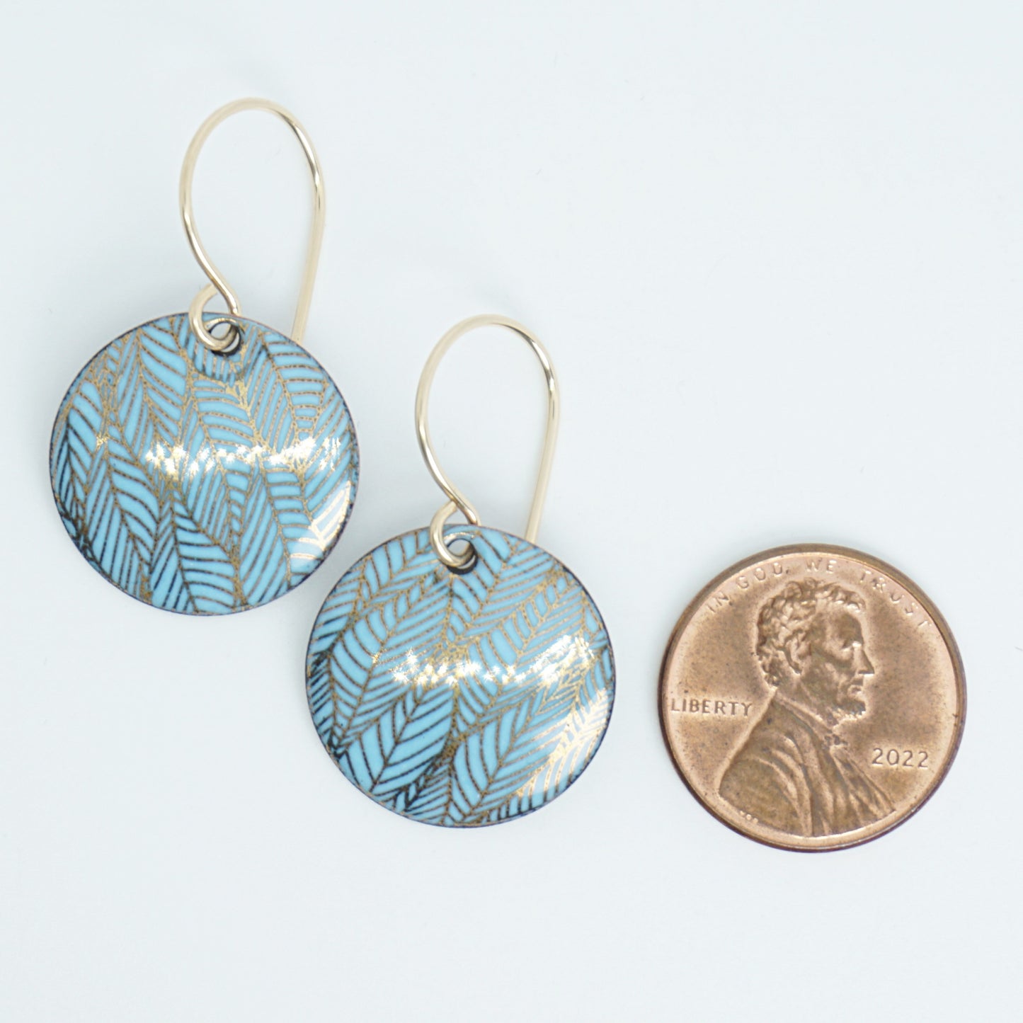 Blue and gold leaf-patterned earrings next to a penny for size comparison on a white background