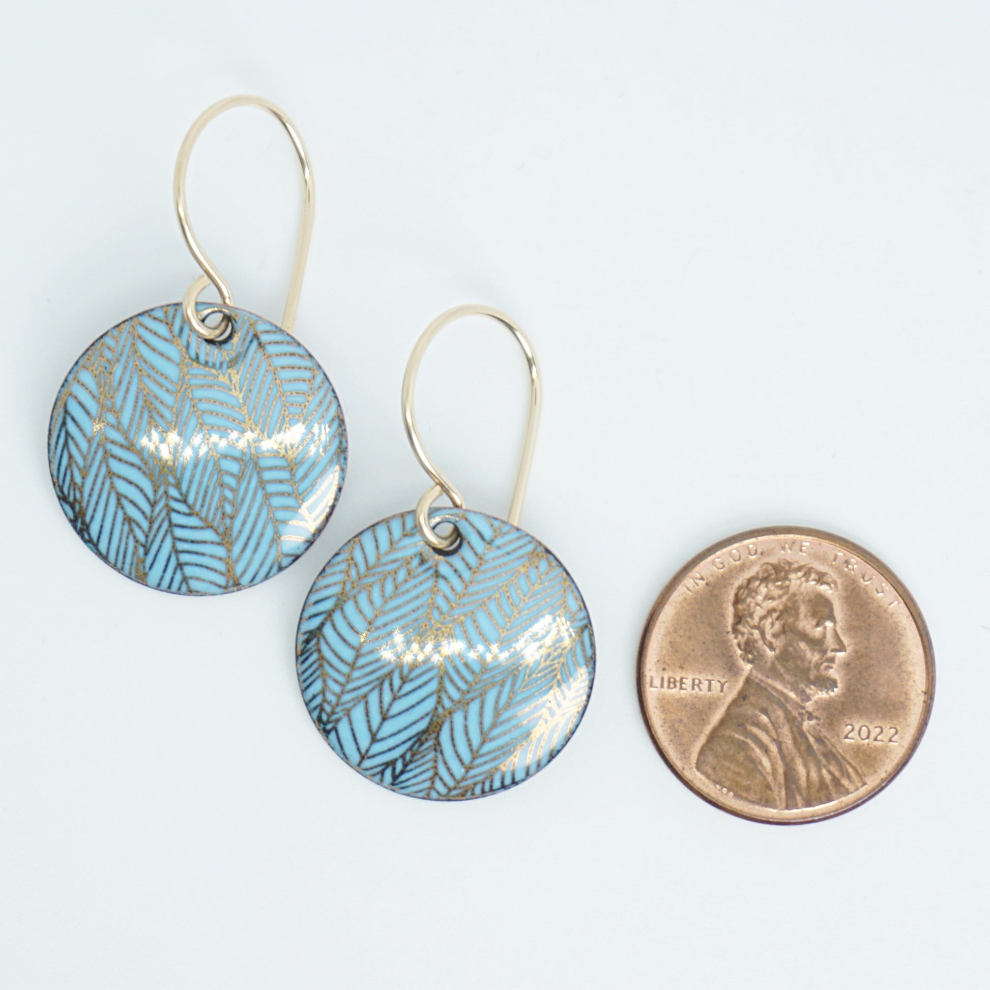 Blue and gold leaf-patterned earrings next to a penny for size comparison on a white background
