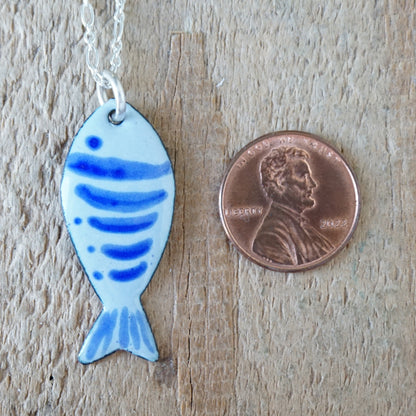 Blue fish-shaped pendant on a chain next to a penny on a wooden surface