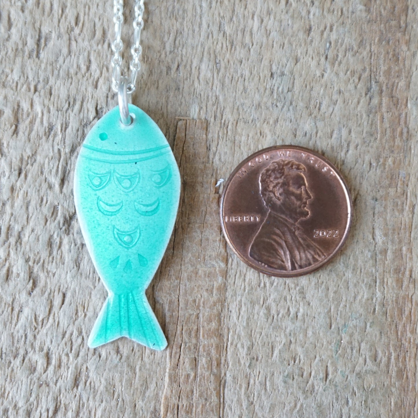 Green fish-shaped pendant on a chain next to a penny for size comparison on a wooden surface.