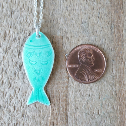 Green fish-shaped pendant on a chain next to a penny for size comparison on a wooden surface.