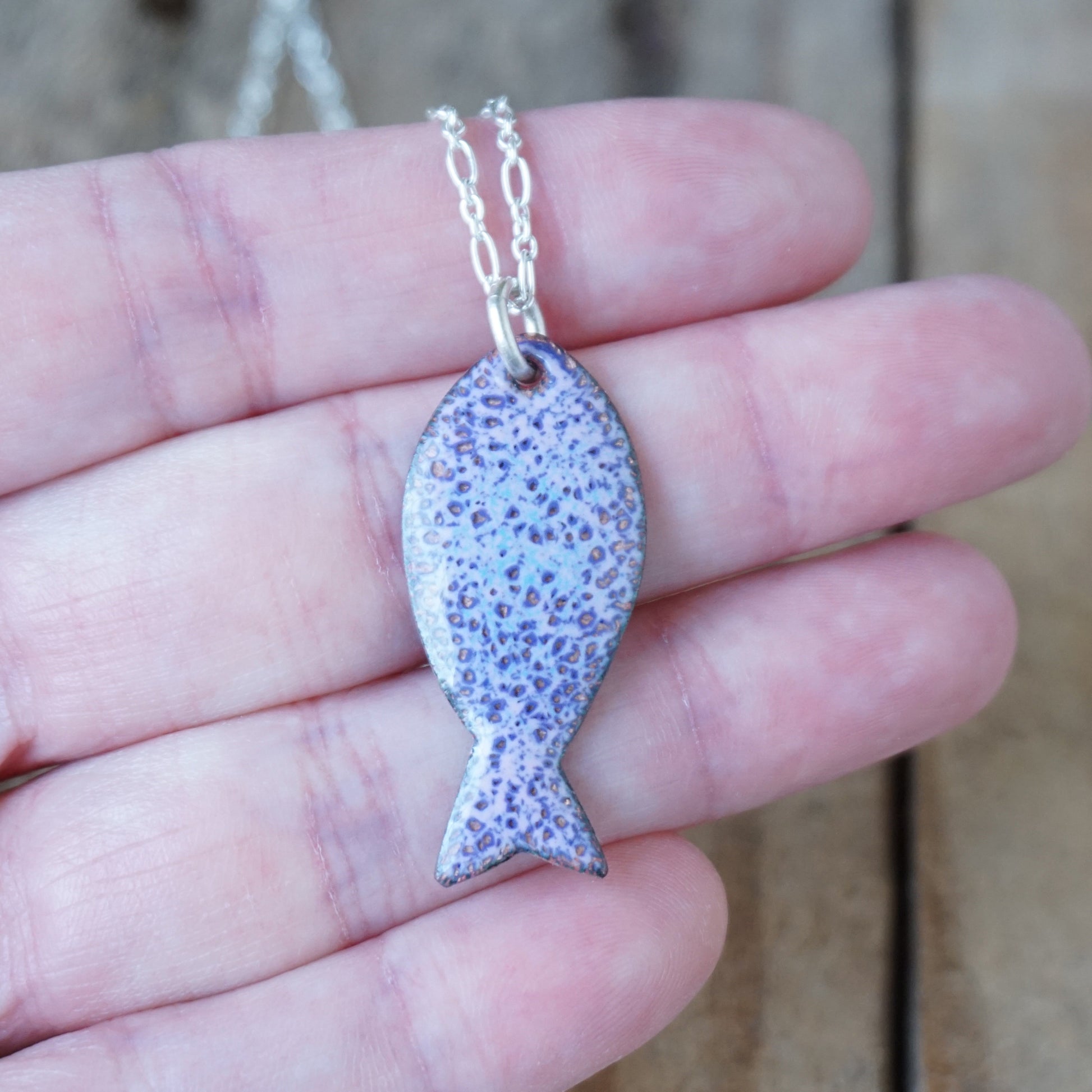 Purple fish-shaped pendant on a chain held in a hand with a blurred background