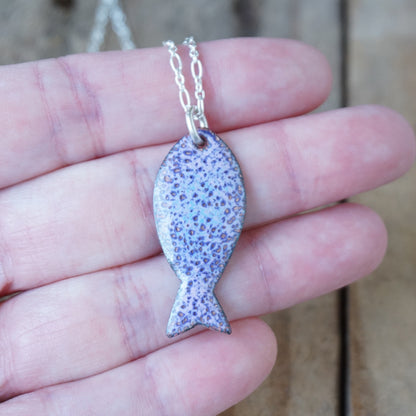 Purple fish-shaped pendant on a chain held in a hand with a blurred background
