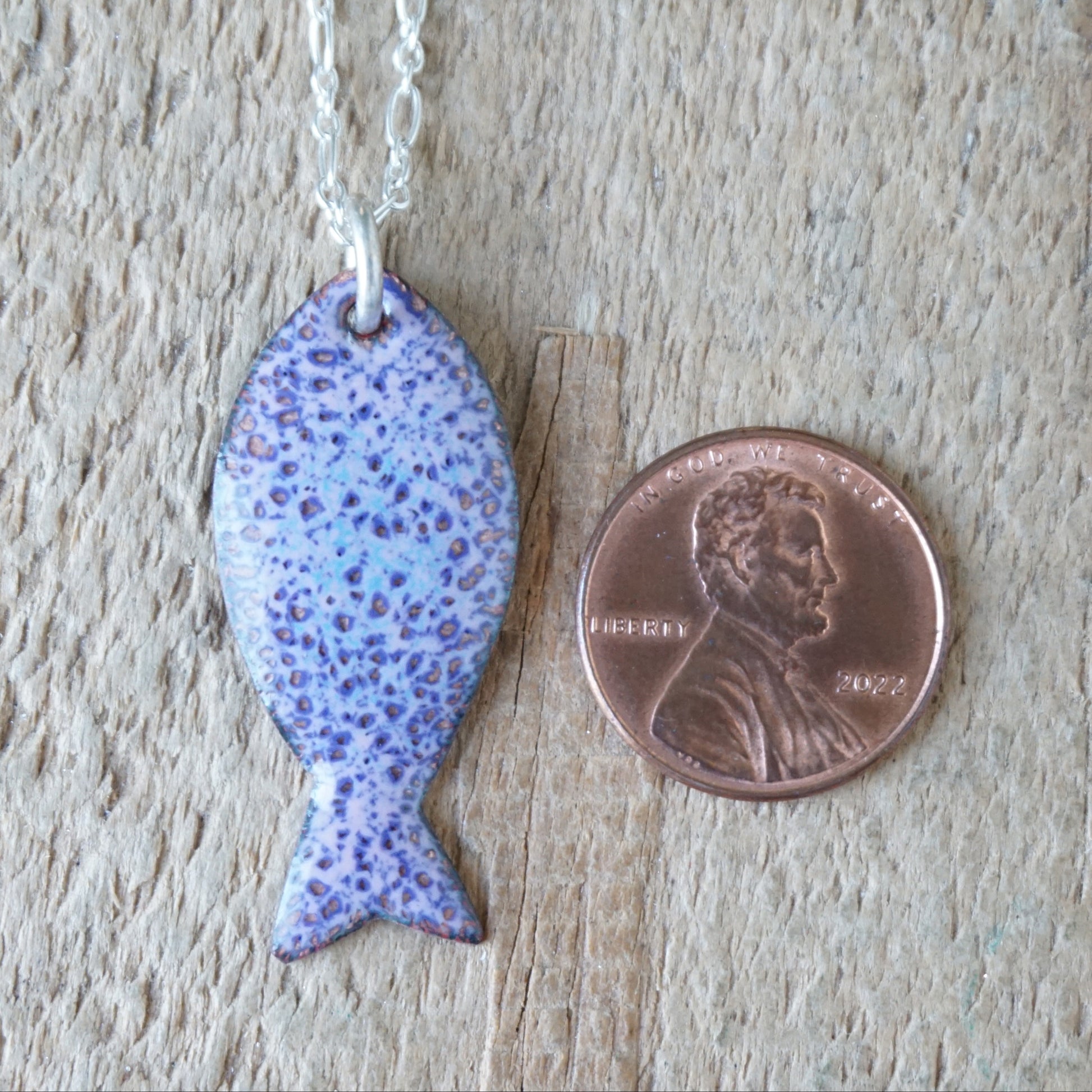 Purple fish-shaped pendant on a chain next to a penny for scale on a wooden surface
