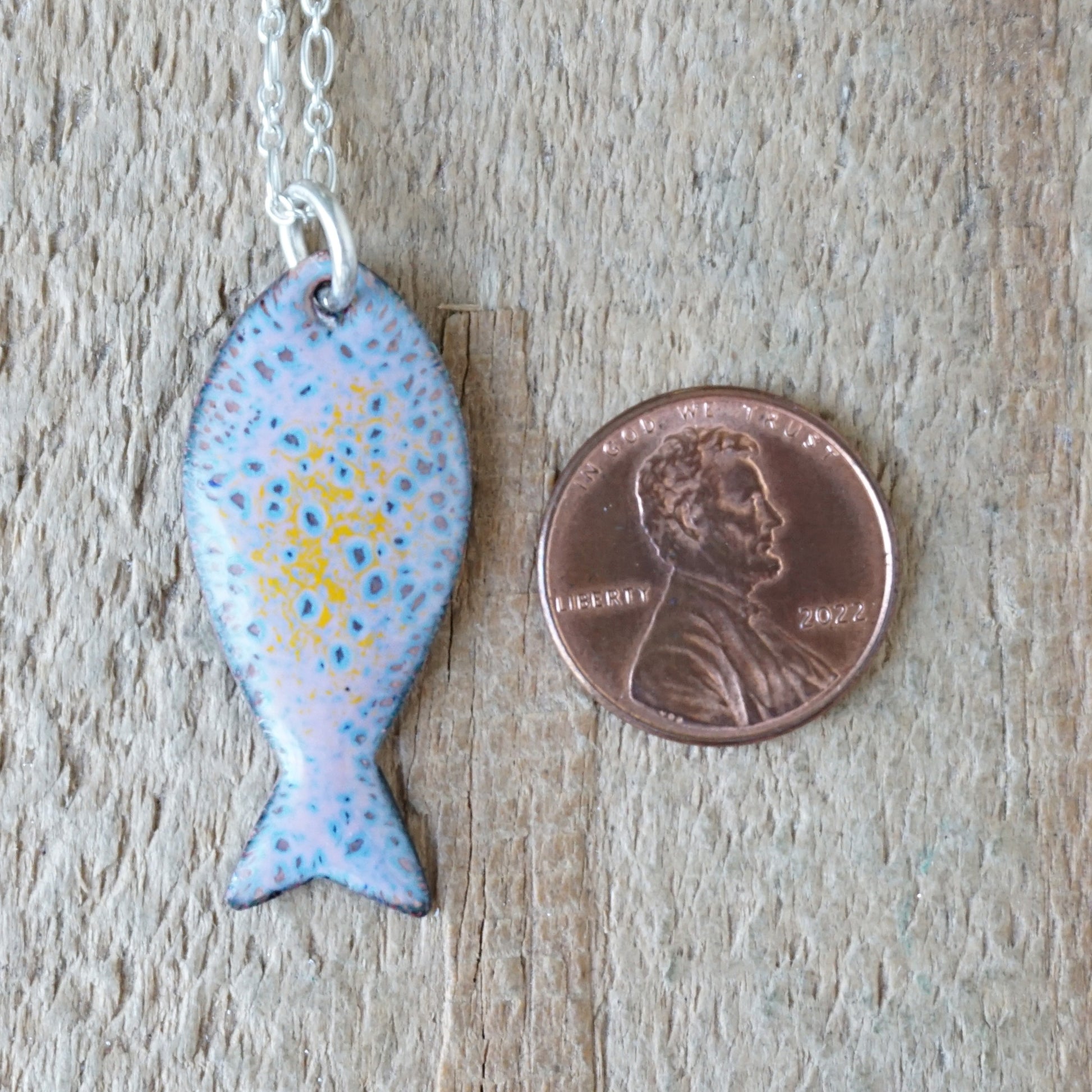 Fish-shaped pendant on a chain next to a penny for scale on a wooden surface