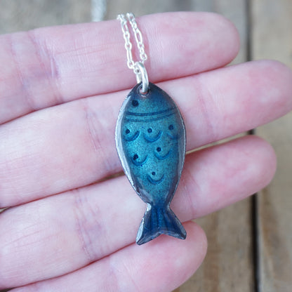 Blue fish-shaped pendant on a chain held in a hand against a wooden background