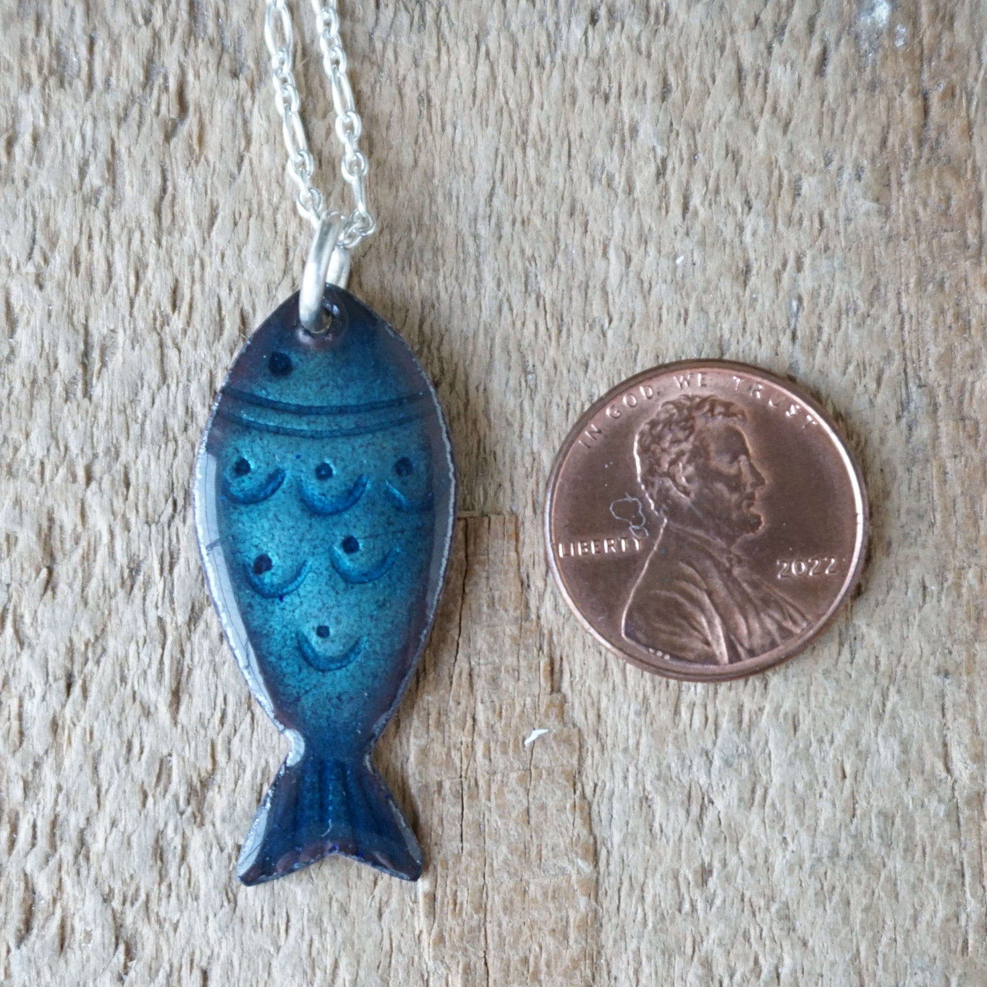 Blue fish-shaped pendant on a chain next to a penny for scale on a wooden surface