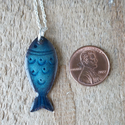 Blue fish-shaped pendant on a chain next to a penny for scale on a wooden surface